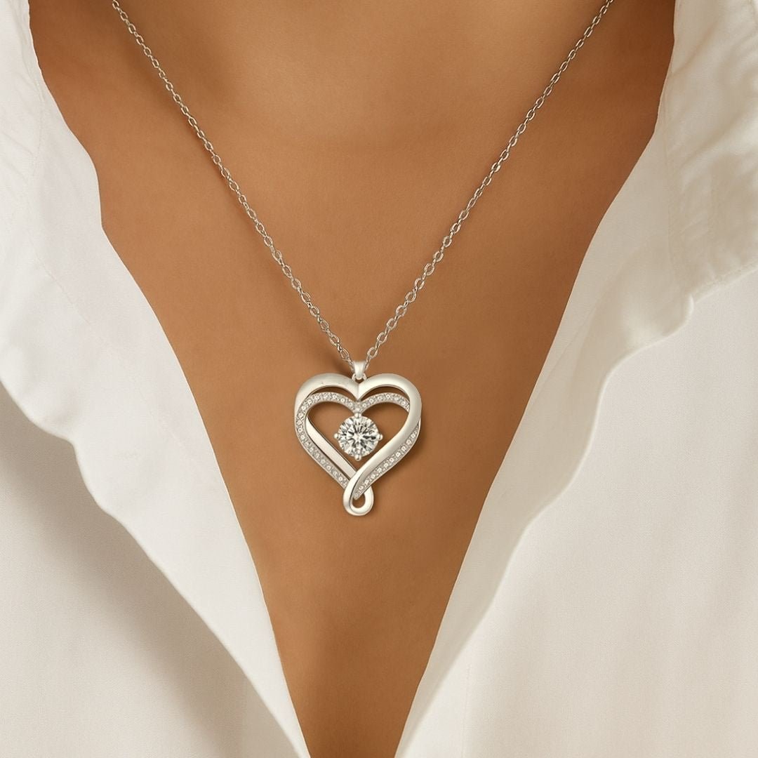 Una persona vestida con una blusa blanca y el "Collar - A mi Esposa", con un colgante plateado en forma de corazón con una gema central brillante, expuesto contra la parte superior desnuda de su pecho, perfecto como regalo parejas.