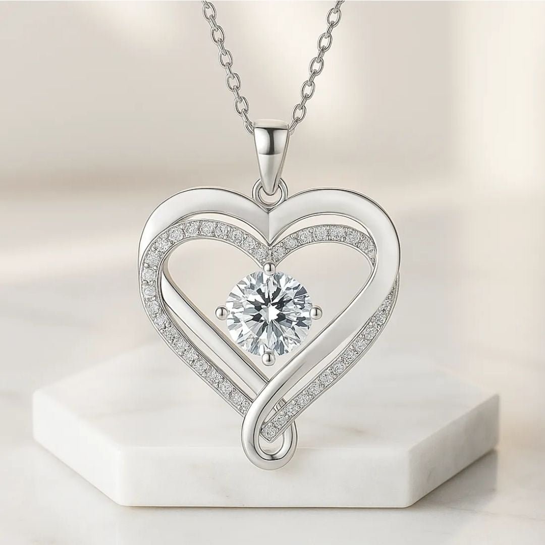 Un "Collar - A mi Esposa" de plata en forma de corazón con pequeños diamantes y un gran diamante central redondo, que se muestra sobre una plataforma hexagonal de mármol-un elegante collar de acero inoxidable, ideal como regalo para parejas.